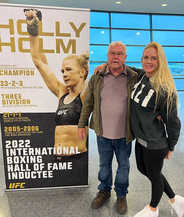 Holly Holm in posa con il padre di fronte alla locandina della sua elezione come membro della International Boxing Hall of Fame - Kombatnet News